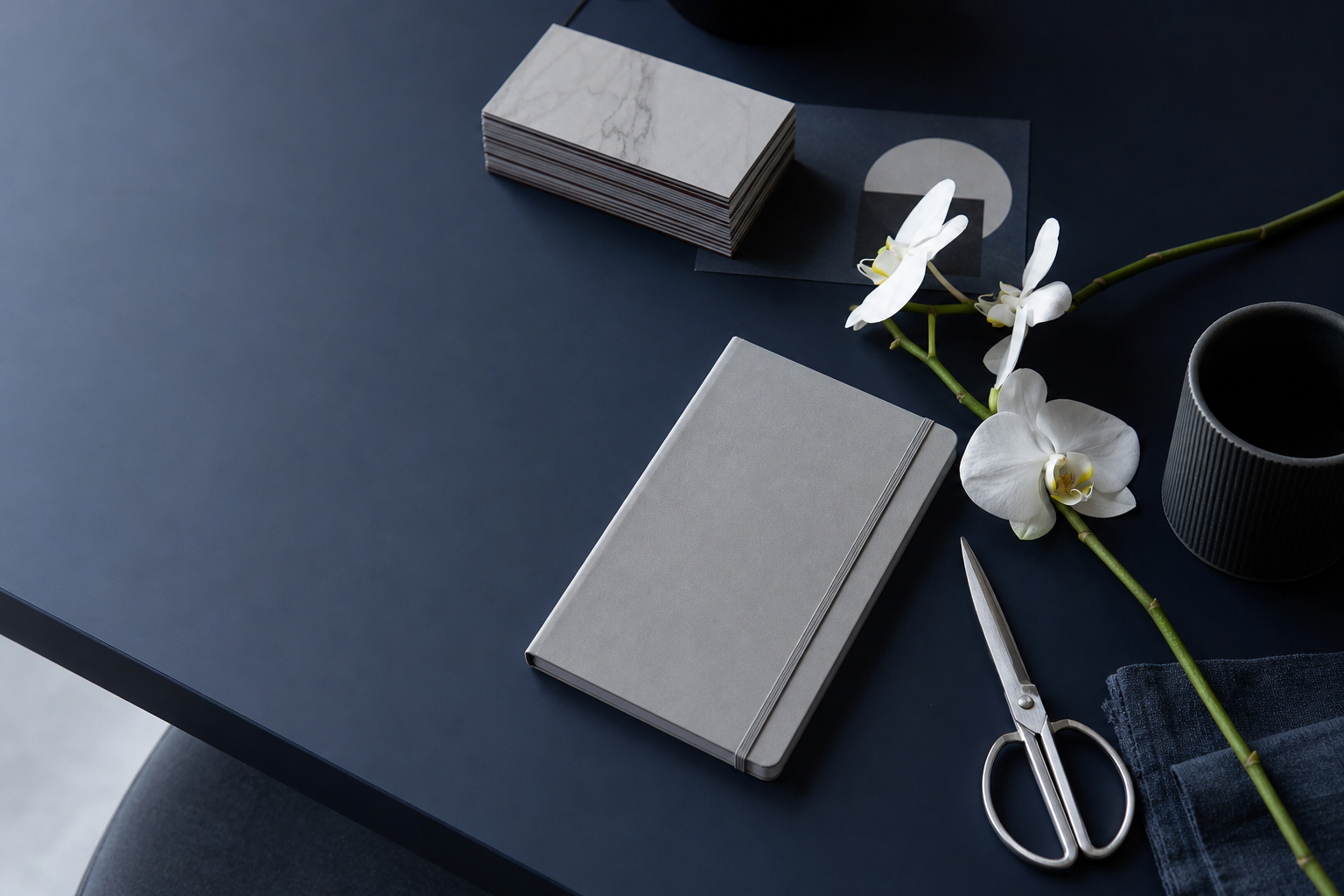 Cool navy studio desk seen from above with a grey notebook, steel scissors and a white orchid